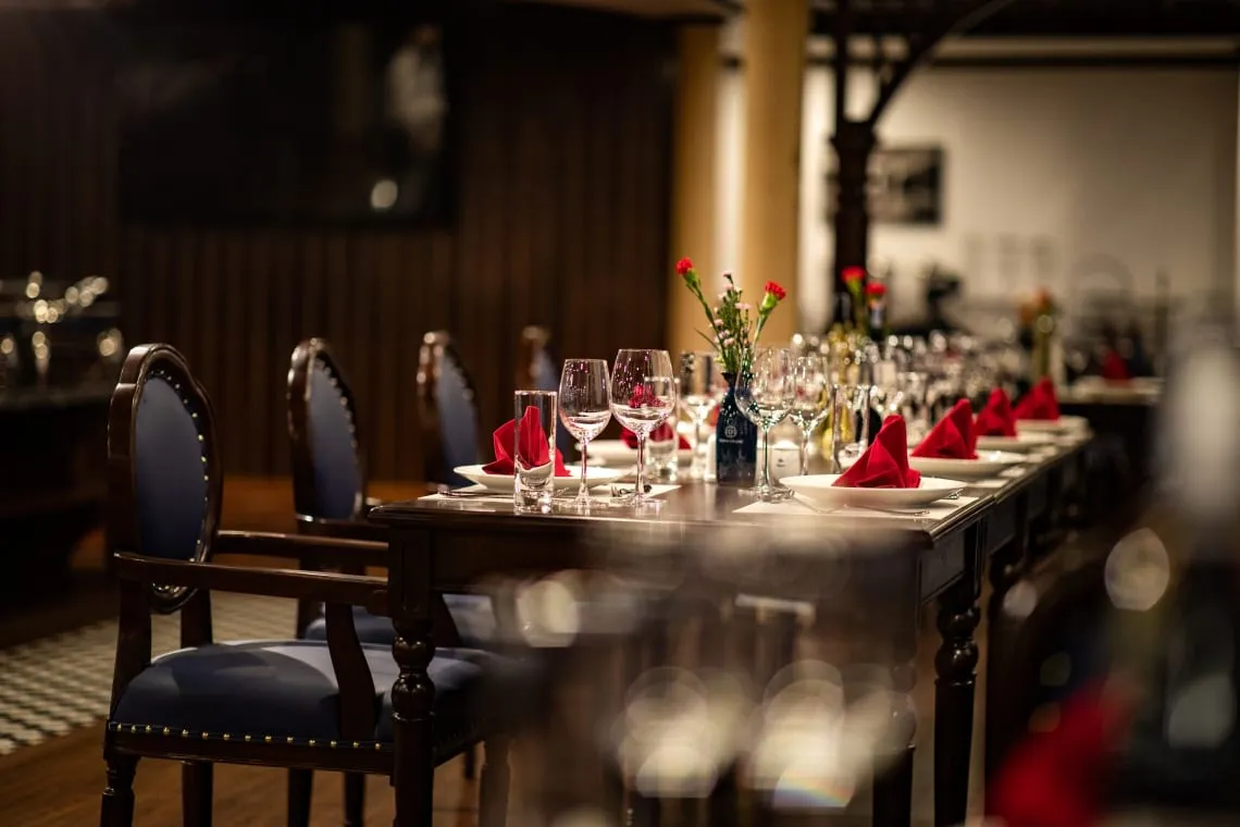 A long dark wooden dining table set with wine glasses, white plates, red folded napkins and small red flowers, flanked by navy upholstered chairs in a dimly lit interior.