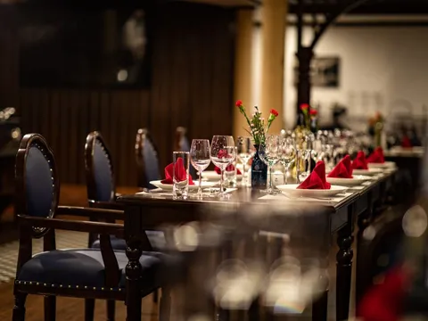 A long dark wooden dining table set with wine glasses, white plates, red folded napkins and small red flowers, flanked by navy upholstered chairs in a dimly lit interior.