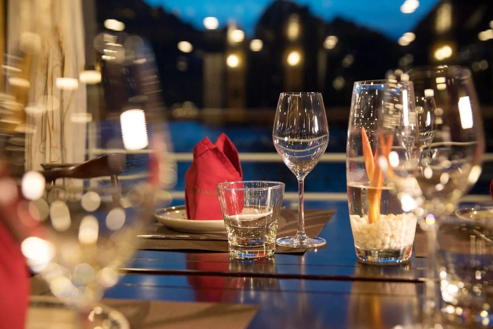 A dusk dining table set with wine glasses, a tumbler, a red folded napkin with faint embroidered text and a vase of white pebbles with an orange flower, in front of blurred lights and dark karst-like silhouettes over water.