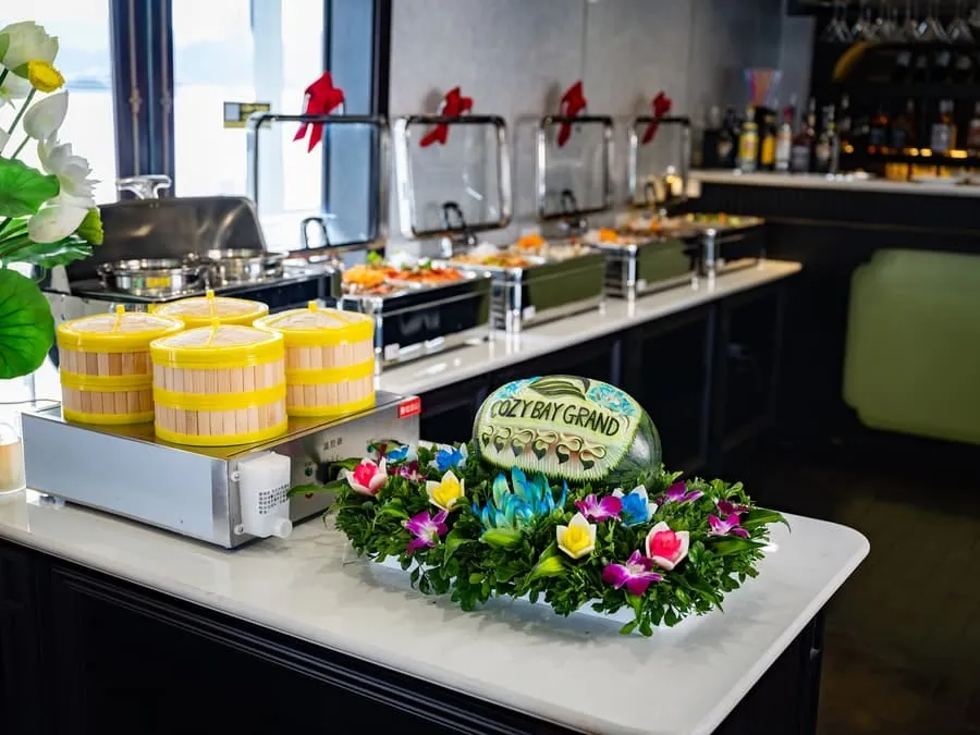 A buffet counter with steamer baskets, chafing dishes, and a carved watermelon reading Cozy Bay Grand.