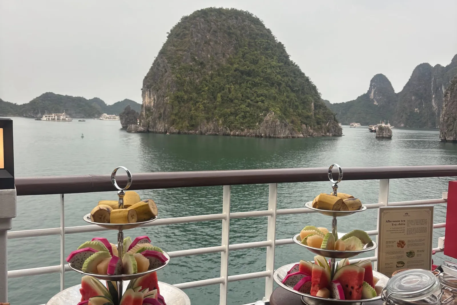 Tiered serving trays with fruit and snacks arranged on a table with karst limestone islands visible through boat deck railing.