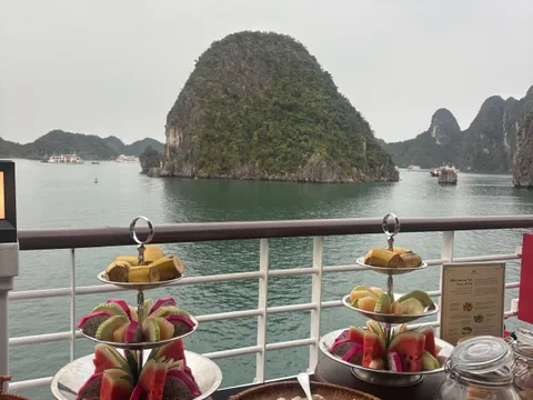 Tiered serving trays with fruit and snacks arranged on a table with karst limestone islands visible through boat deck railing.