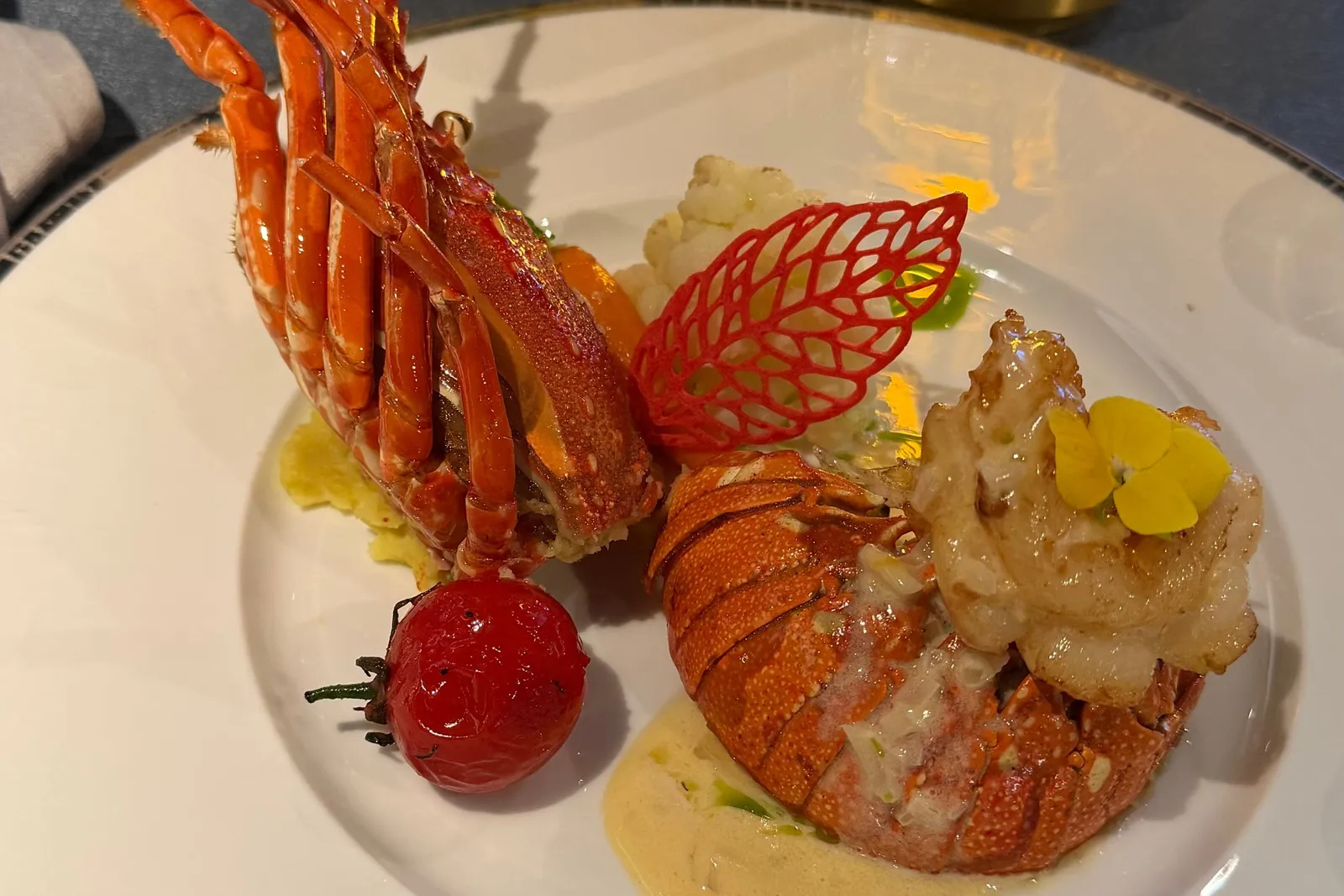 A plated lobster dish with a red tuile leaf, cherry tomato, cauliflower, cream sauce and a yellow flower on a white rimmed plate, set on a dining table with a glowing gold lamp, vase and salt and pepper shakers.