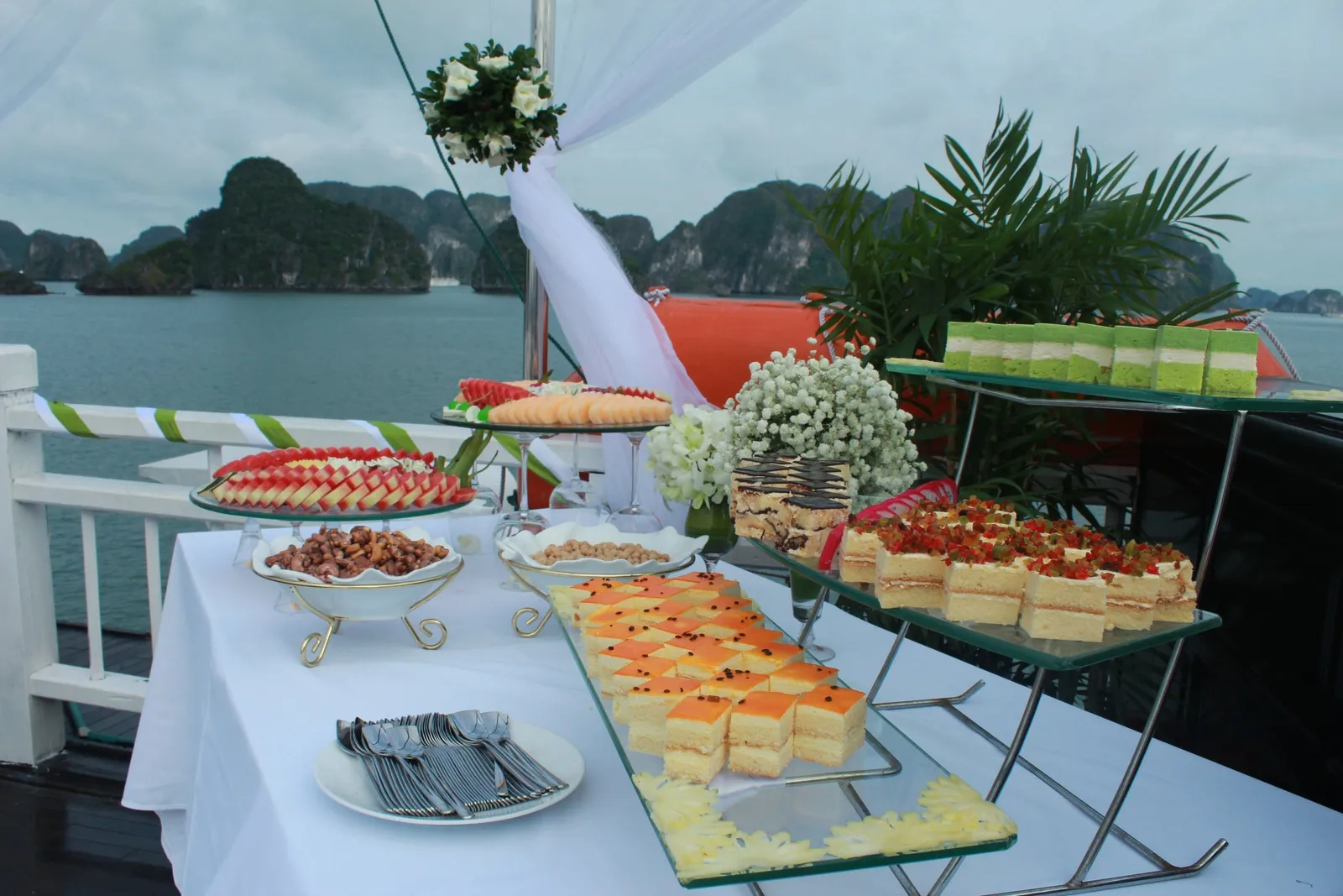 A buffet table with desserts and appetizers is set up on a boat deck with limestone karsts visible across the water in the background.