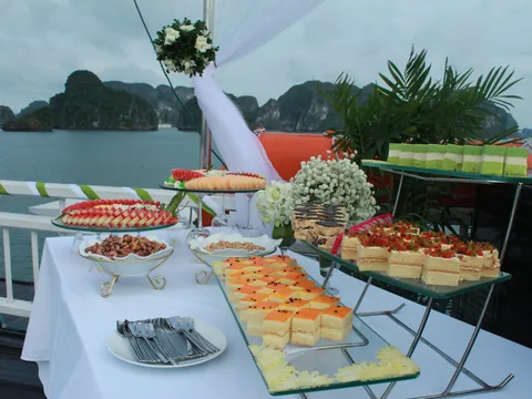 A buffet table with desserts and appetizers is set up on a boat deck with limestone karsts visible across the water in the background.