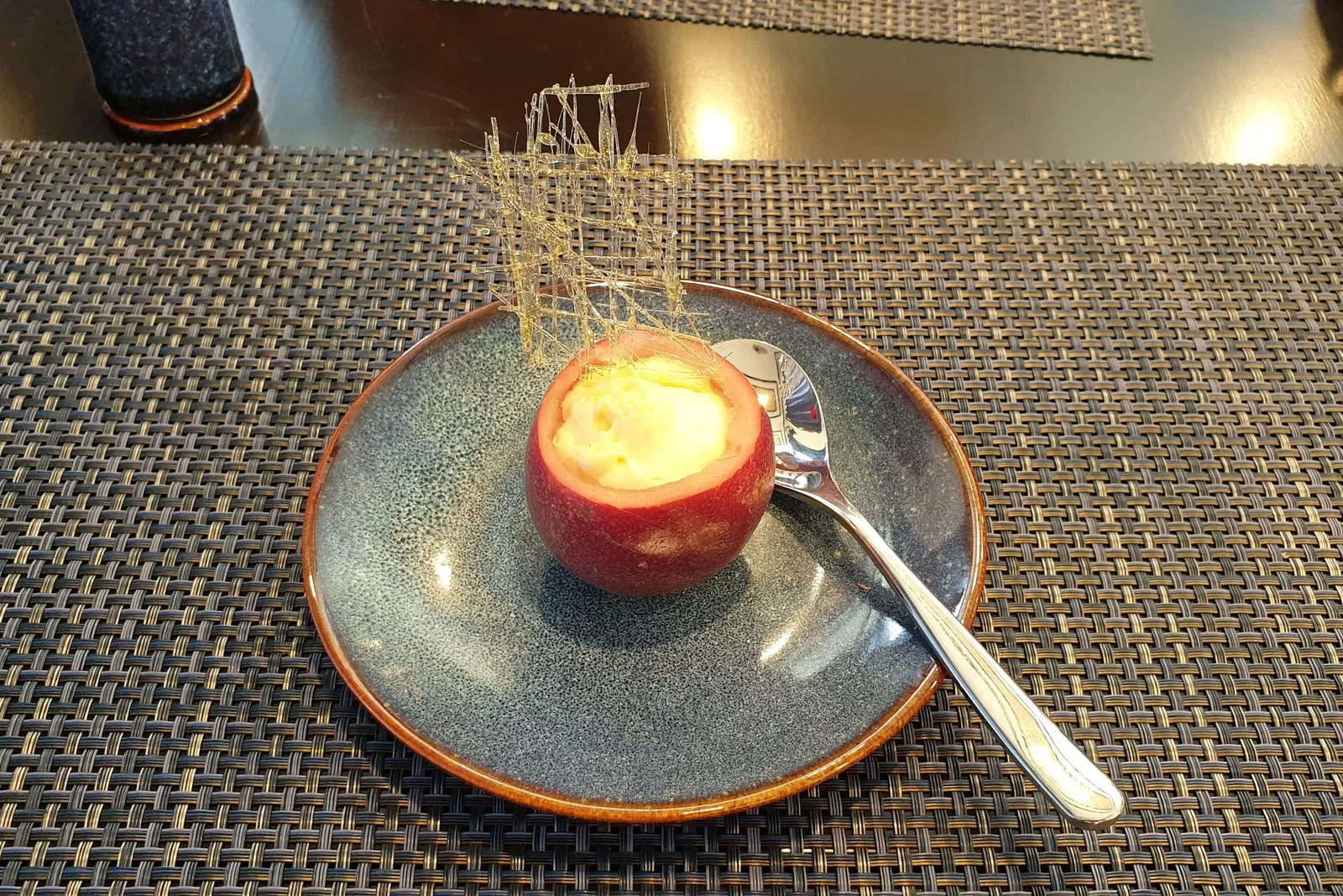 A plated dessert served in a hollowed tomato with a decorative garnish on a woven placemat at a dining table.