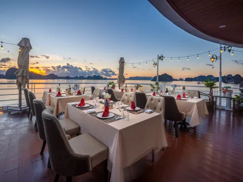 An open-air dining deck at sunset with tables set with white linen and red napkins, string lights and a view of limestone islands.