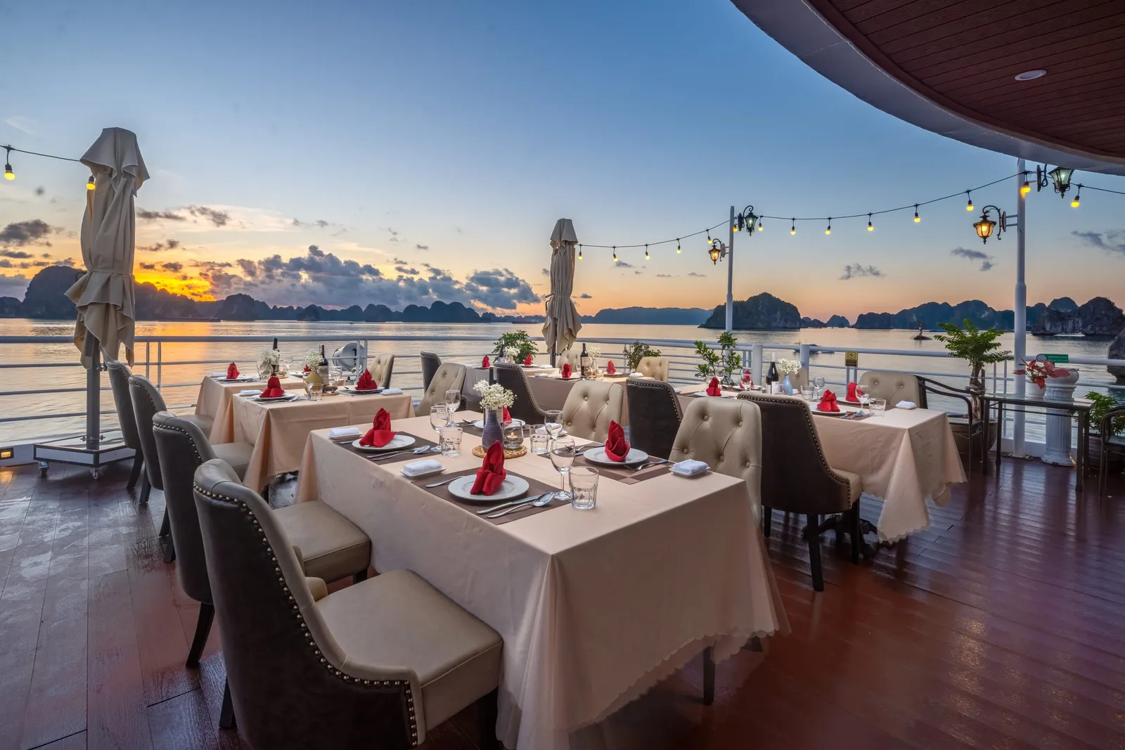 An open-air dining deck at sunset with tables set with white linen and red napkins, string lights and a view of limestone islands.