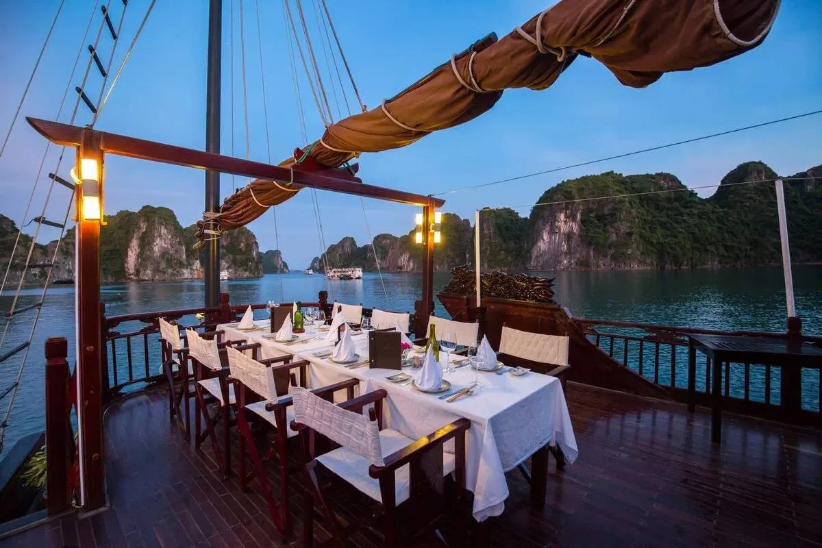 A long table set with white linen on the open rear deck at dusk, with karst islands beyond the railing.