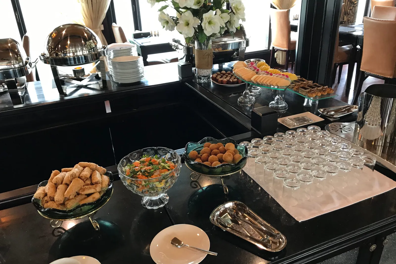 A buffet table with pastries, salads, and serving dishes set up in a dining room with large windows and curtains.