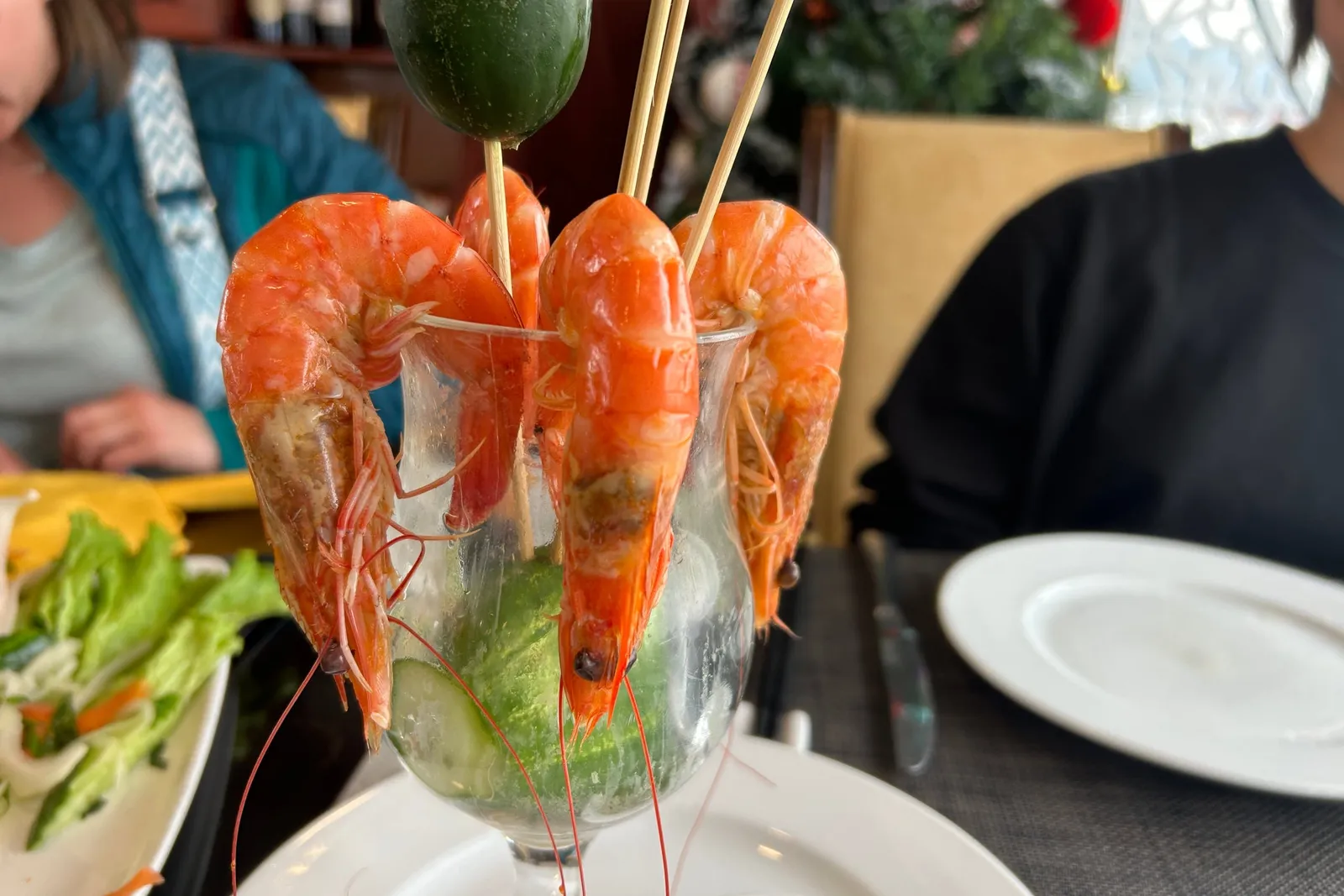 A glass containing cooked shrimp on skewers with cucumber and vegetables sits on a dining table with people seated around it.