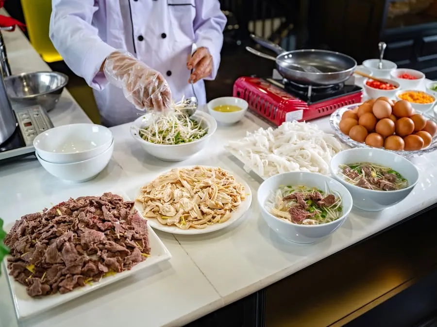 A pho station with sliced beef, chicken, noodles, herbs and eggs laid out, with a chef preparing bowls.