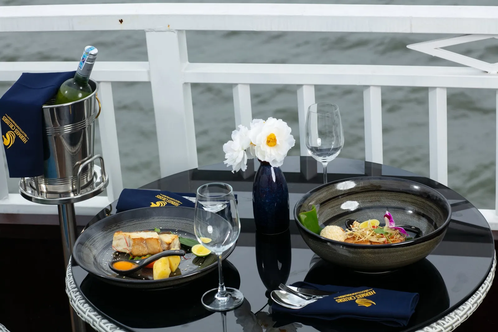A black round table on a white-railed deck set with two plated dishes, wine glasses, a white flower vase and a wine bucket, with navy napkins embroidered 'Hermes Cruises' and grey water behind.