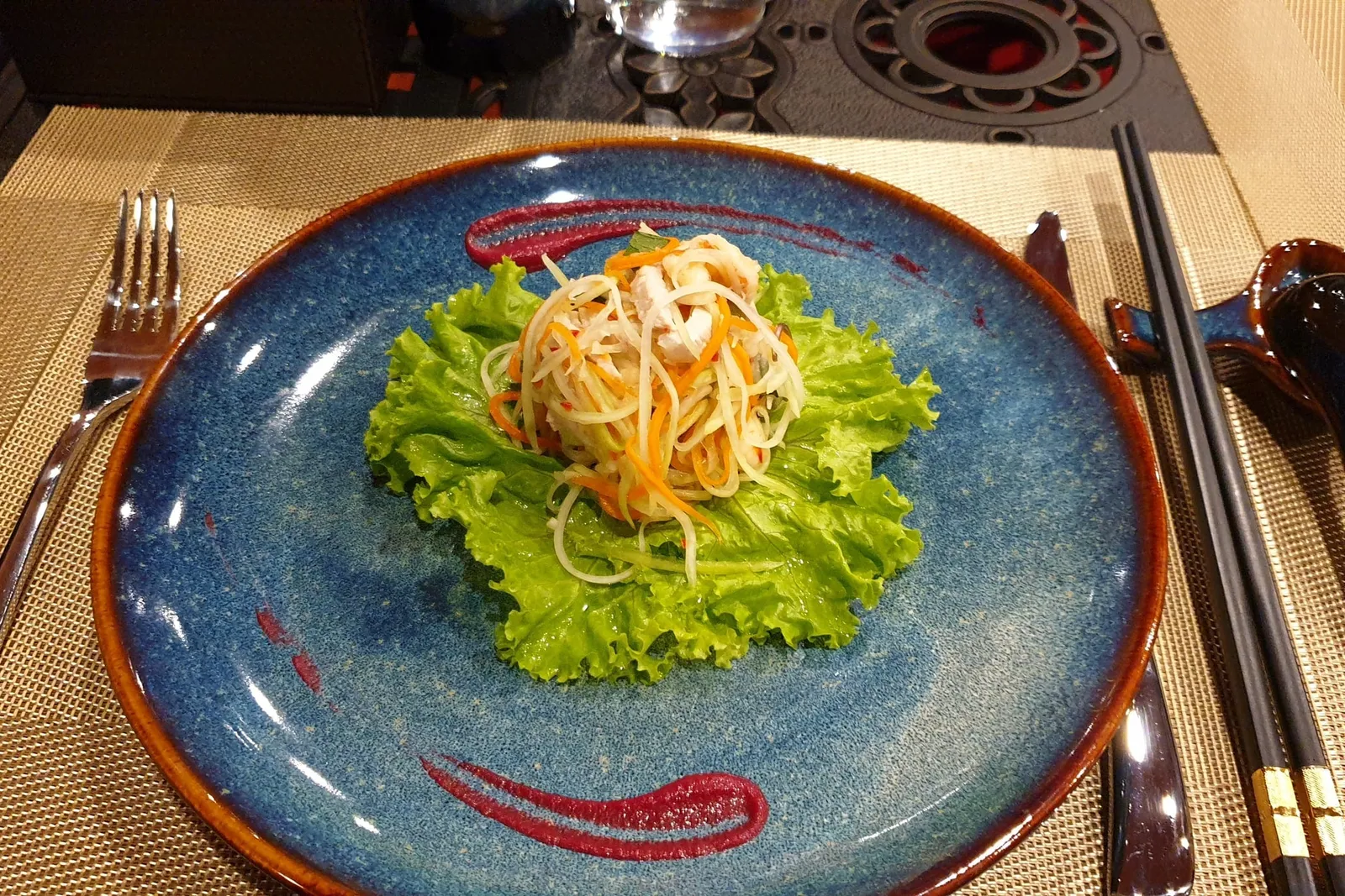 A plated salad dish with noodles on lettuce sits on a blue ceramic plate with chopsticks, fork, and spoon on a woven placemat.