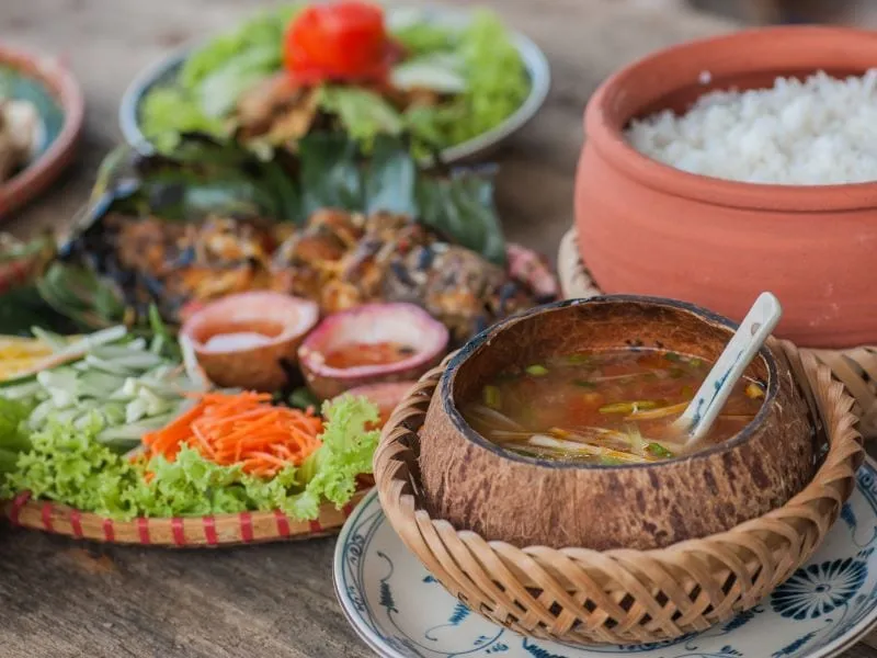 A Vietnamese lunch spread with grilled fish, vegetables, rice in a clay pot and soup in a coconut bowl.