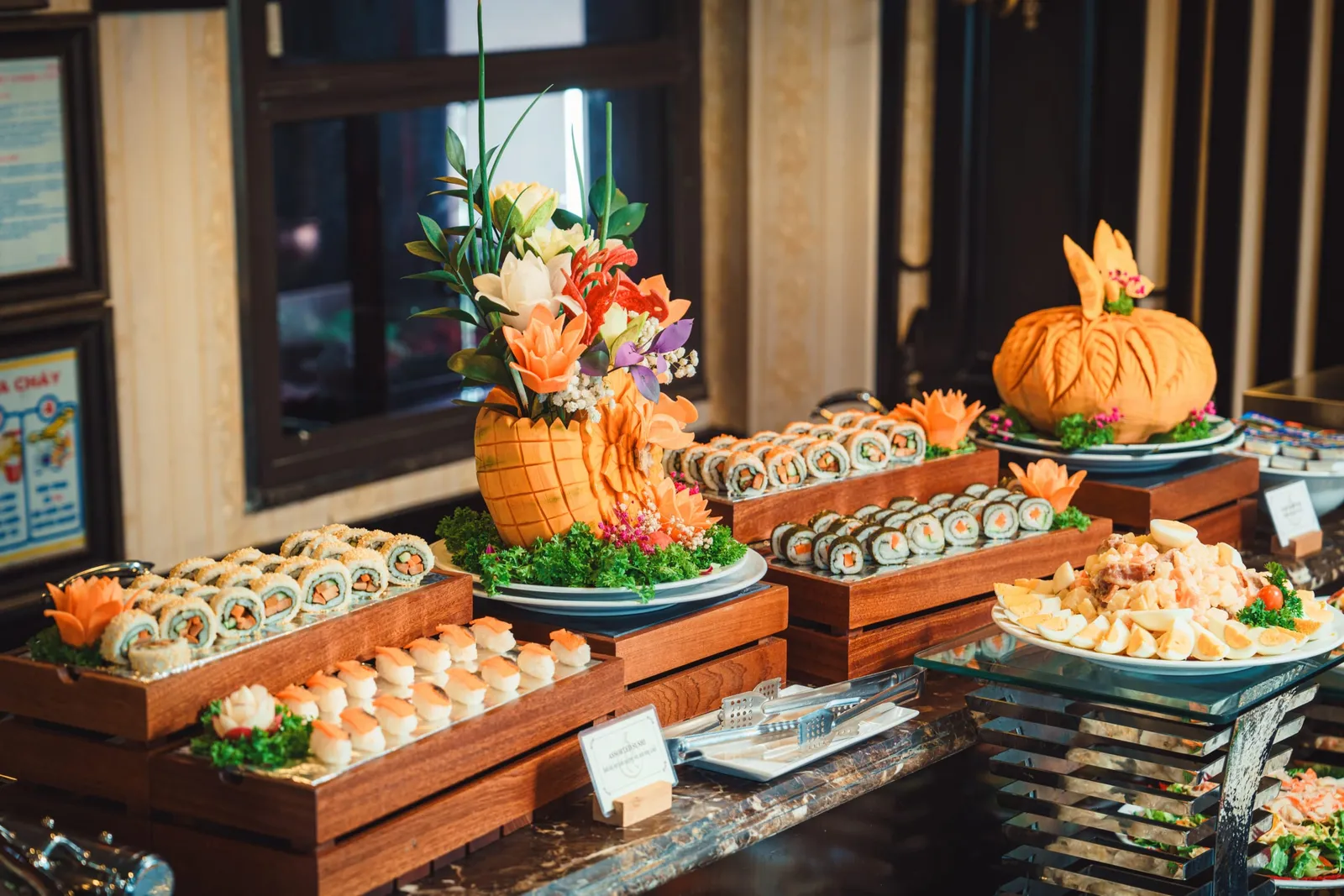 An indoor buffet counter with sushi rolls, nigiri, egg salad and carved fruit and flower centerpieces on wooden risers, with a window and Vietnamese-language wall notices behind.