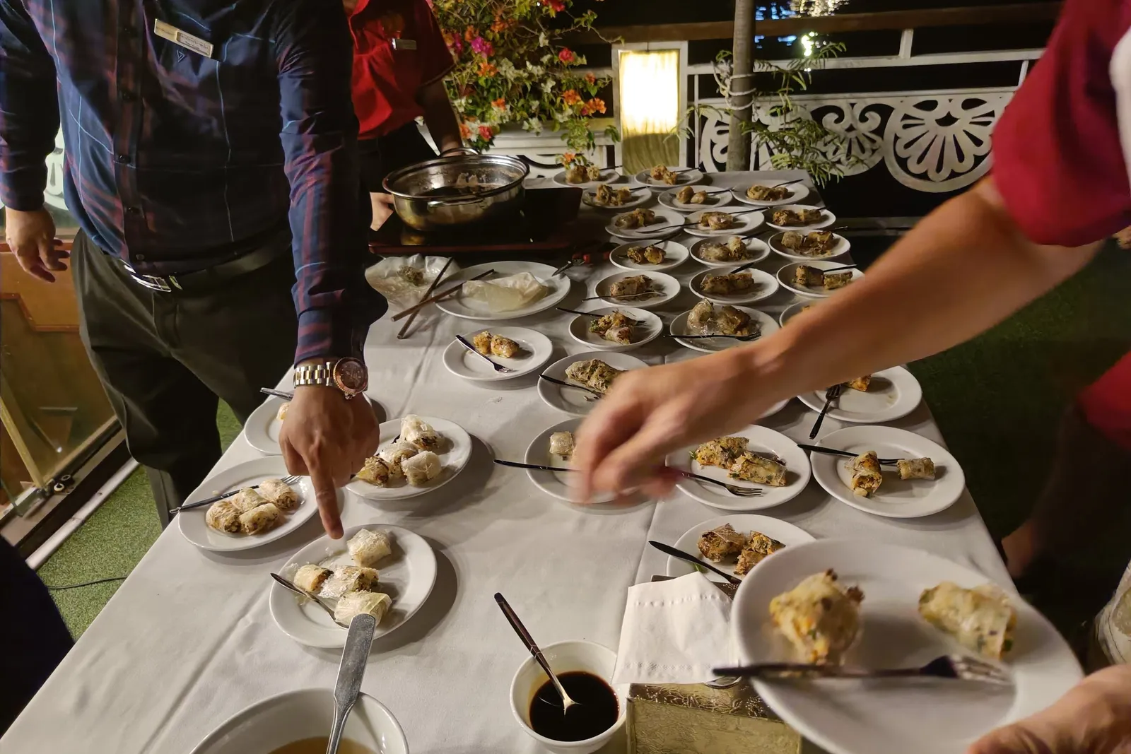 Staff members plating food at a buffet table on a boat deck at night with decorative railings and flowers visible in the background.
