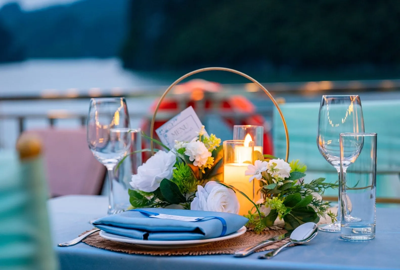 A dusk dinner table on a deck with a blue tablecloth, folded blue napkin, wine glasses, a lit candle and a white floral centerpiece with a menu card, with blurred karst hills and water behind.