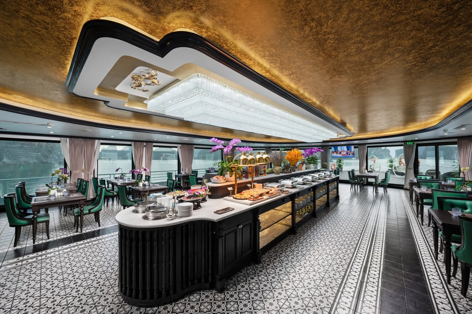 An ornate dining room on a cruise boat with a buffet counter displaying food, green upholstered chairs at tables, panoramic windows showing water and karst landscapes, and an elaborate ceiling with chandelier and gold detailing.