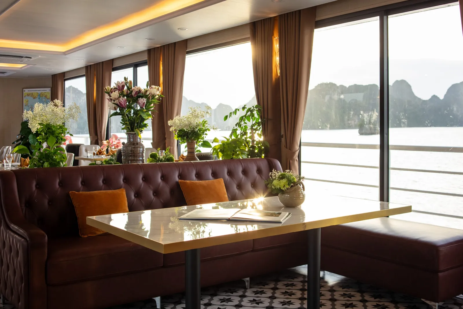 A lounge corner with a tufted brown leather banquette, a marble-top table and fresh flowers beside floor-to-ceiling windows on the bay.