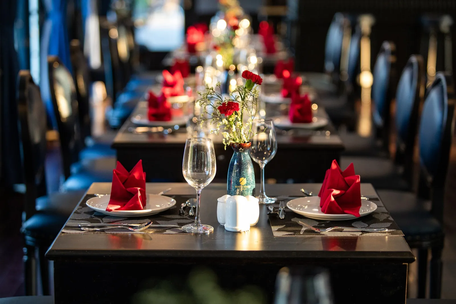Row of dark wooden dining tables set with red folded napkins, wine glasses, and a small vase of red carnations, with dark upholstered chairs in a dimly lit restaurant.