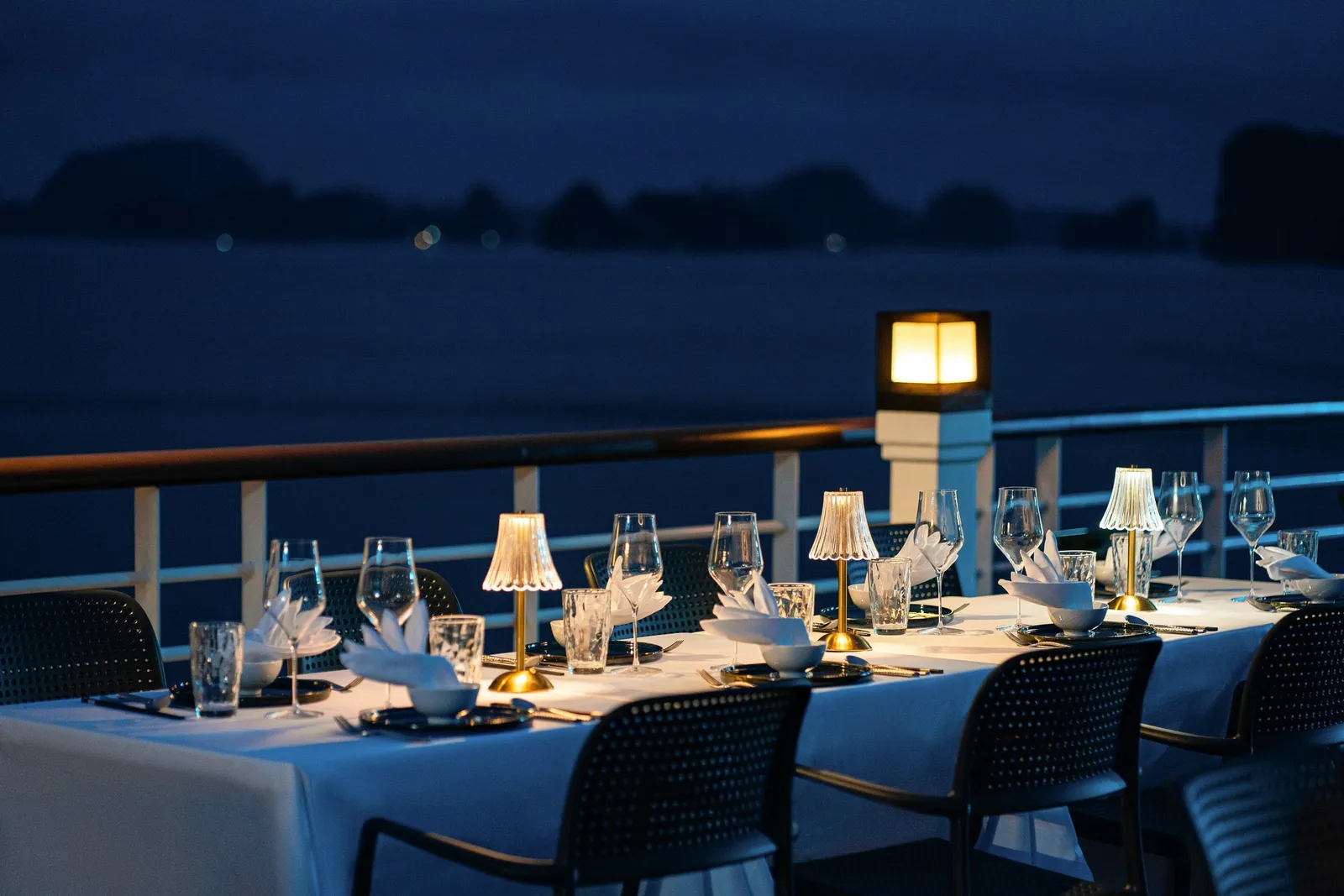 A long dining table set with white linen, glassware, folded napkins and small lamps on an outdoor deck at dusk, with dark silhouetted karst islands across the water.