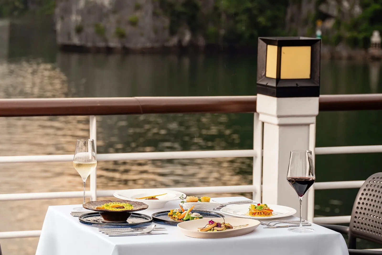 A white-clothed dining table set with several plated dishes and two wine glasses on a railed deck, with calm water and a forested limestone cliff behind.