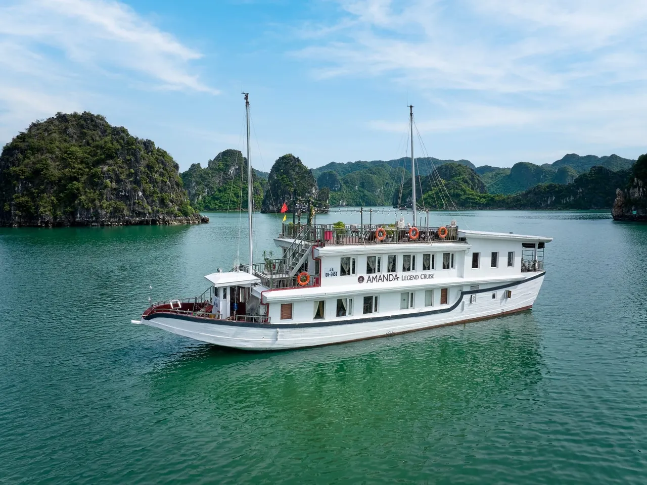 A white two-deck cruise boat with 'Amanda Legend Cruise' lettering on its side floats on green water in front of forested limestone karst islands, photographed from an elevated outside vantage point.