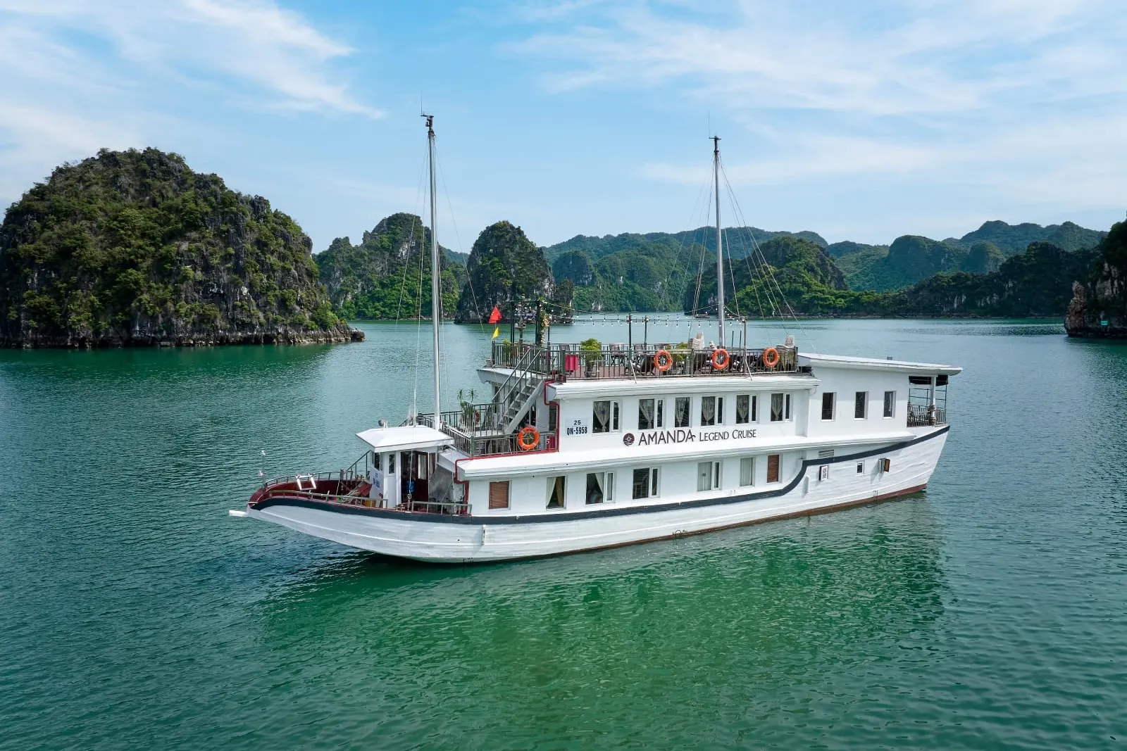 A white two-deck cruise boat with 'Amanda Legend Cruise' lettering on its side floats on green water in front of forested limestone karst islands, photographed from an elevated outside vantage point.