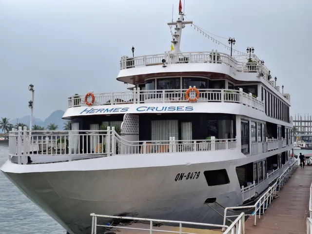 A white multi-deck steel cruise ship moored at a pontoon dock, with 'Hermes Cruises' painted on the upper hull and QN-8479 on the bow, under a hazy sky with karst hills in the distance.