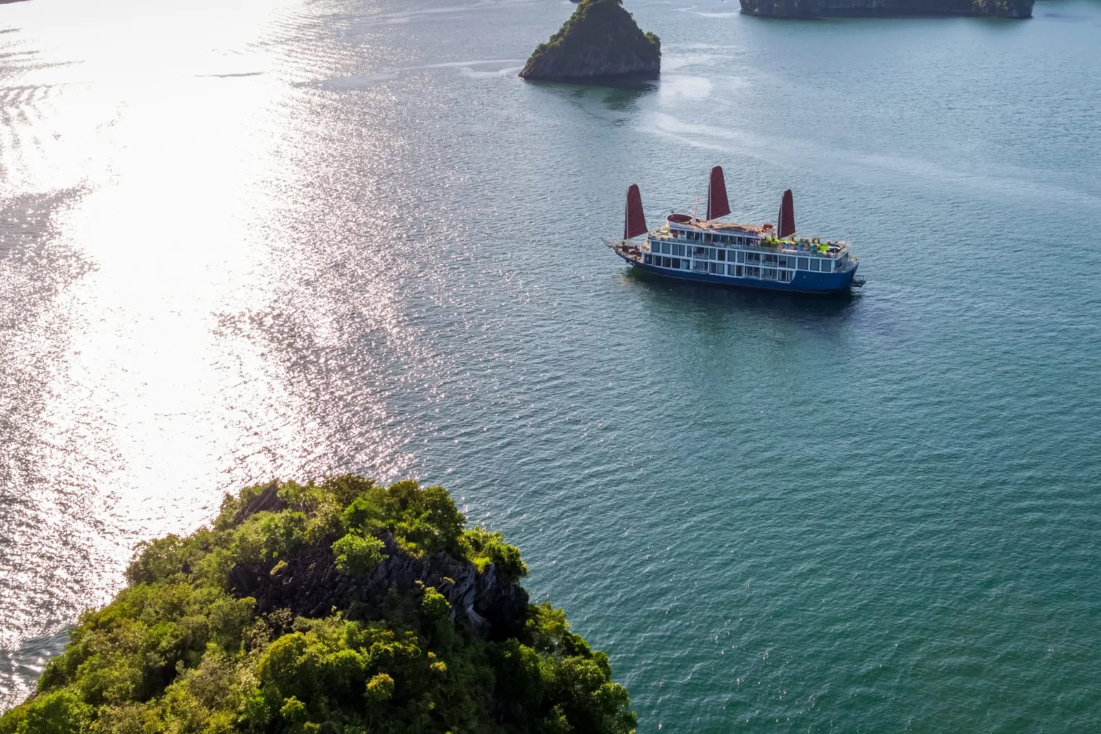An aerial view of a blue-hulled cruise boat with three orange sails on sun-glittered water in front of limestone karst islands.