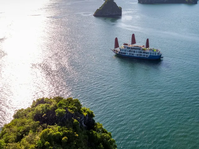 An aerial view of a blue-hulled cruise boat with three orange sails on sun-glittered water in front of limestone karst islands.