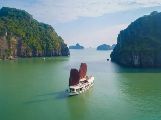 An aerial view of a white junk-style boat with two dark red sails on green water between forested limestone karst cliffs, with more karsts in the hazy distance.