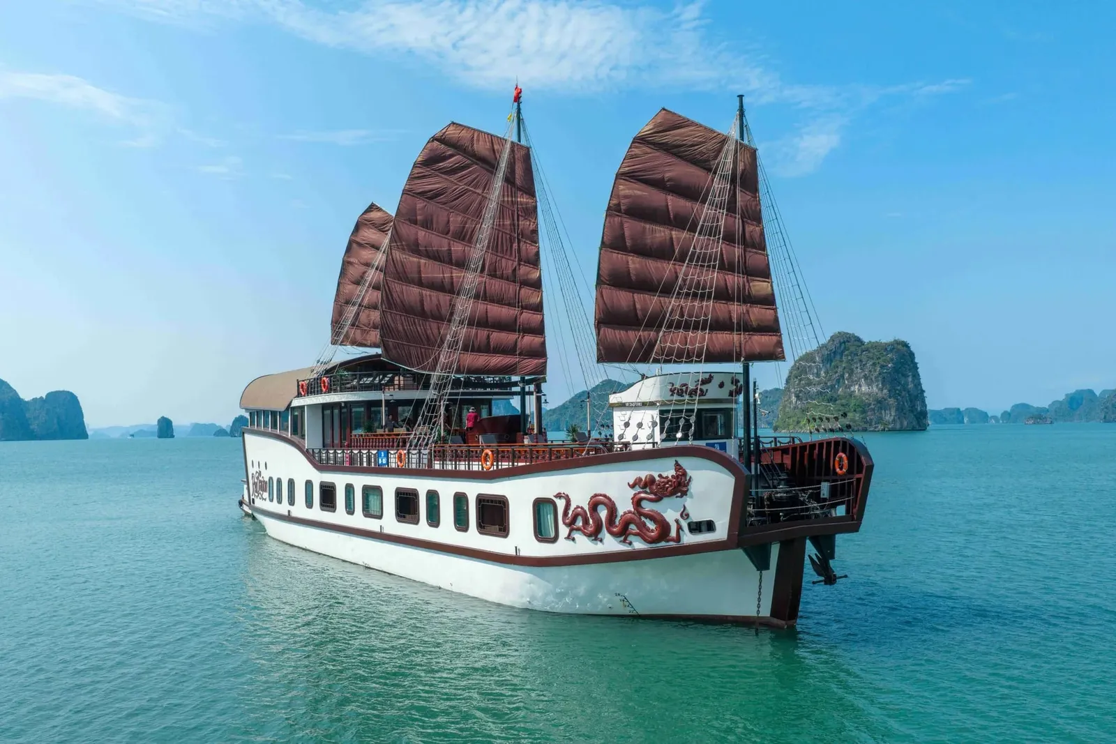 A white junk-style cruise boat with brown sails and a red dragon design on the hull sits on calm green water in front of limestone karst islands under a blue sky.