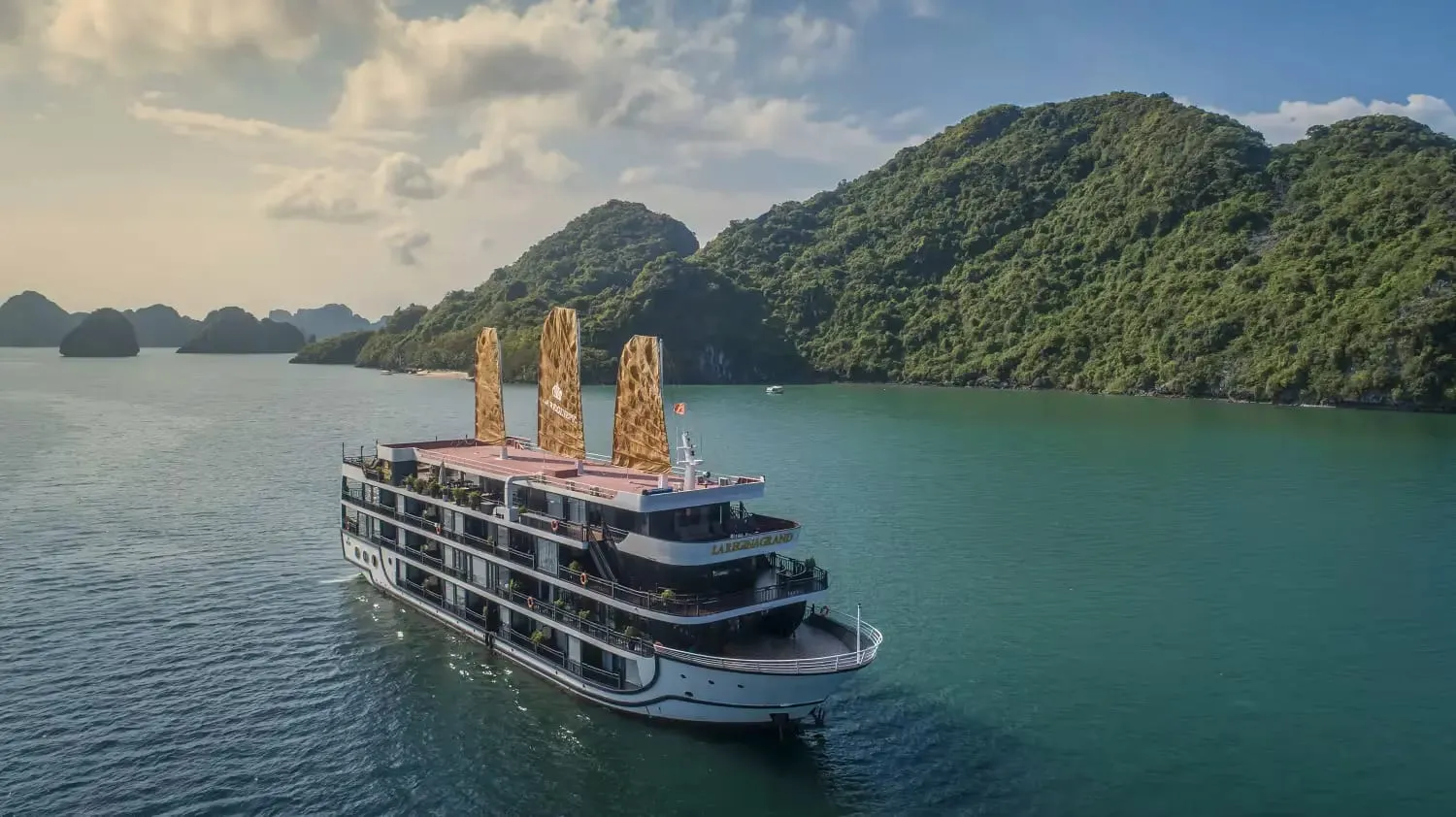 Aerial view of a multi-deck white cruise ship with golden junk-style sails and name lettering on its stern, floating on green water beside forested limestone islands under a partly cloudy sky.