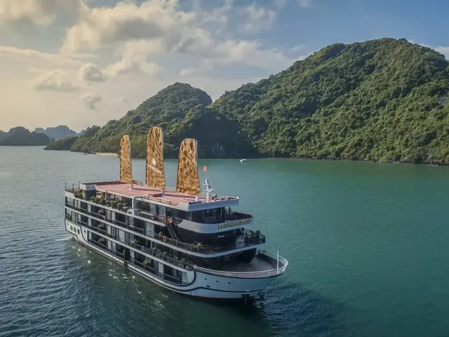 Aerial view of a multi-deck white cruise ship with golden junk-style sails and name lettering on its stern, floating on green water beside forested limestone islands under a partly cloudy sky.