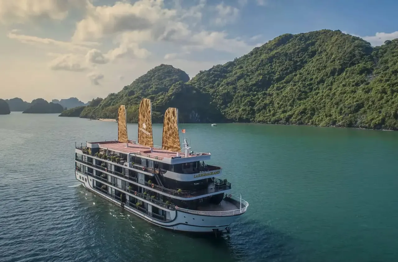 Aerial view of a multi-deck white cruise ship with golden junk-style sails and name lettering on its stern, floating on green water beside forested limestone islands under a partly cloudy sky.