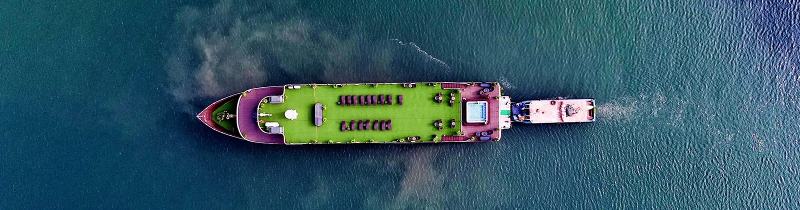 Top-down aerial view of a large vessel with a green-covered upper deck, loungers and a small pool, towing or accompanied by a smaller boat on teal water with a churned wake.