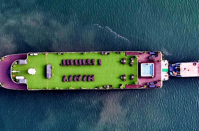 Top-down aerial view of a large vessel with a green-covered upper deck, loungers and a small pool, towing or accompanied by a smaller boat on teal water with a churned wake.