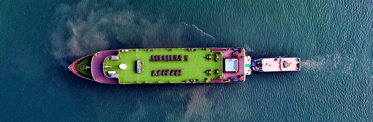 Top-down aerial view of a large vessel with a green-covered upper deck, loungers and a small pool, towing or accompanied by a smaller boat on teal water with a churned wake.