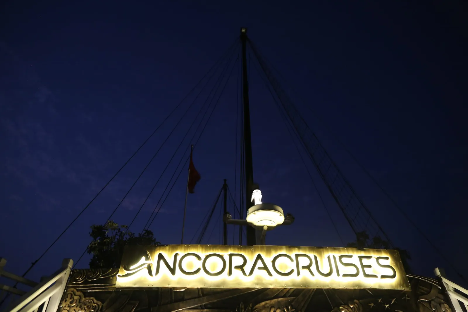 An illuminated sign reading ANCORACRUISES mounted on a boat's structure at night with a lamp and flag above it.