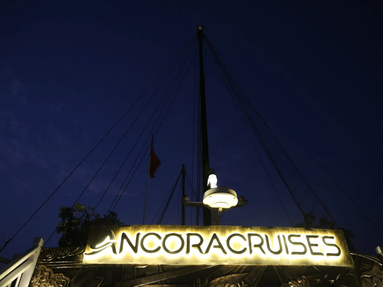 An illuminated sign reading ANCORACRUISES mounted on a boat's structure at night with a lamp and flag above it.
