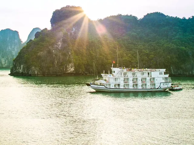 A white multi-deck cruise boat with a small script name on its hull sits on calm green water in front of forested limestone karst cliffs, with the sun flaring above the ridge.