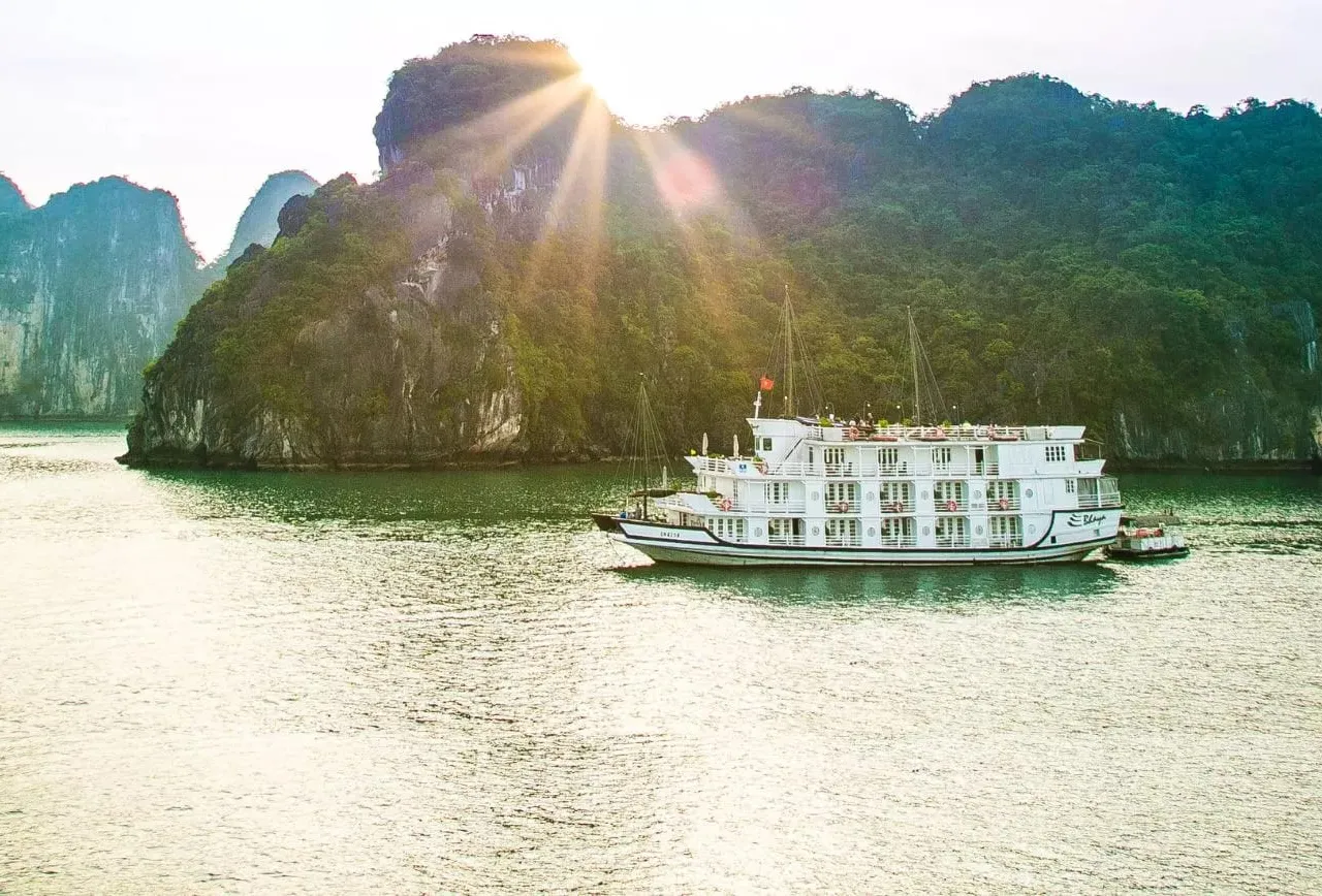A white multi-deck cruise boat with a small script name on its hull sits on calm green water in front of forested limestone karst cliffs, with the sun flaring above the ridge.