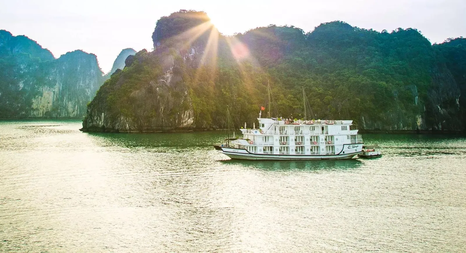 A white multi-deck cruise boat with a small script name on its hull sits on calm green water in front of forested limestone karst cliffs, with the sun flaring above the ridge.