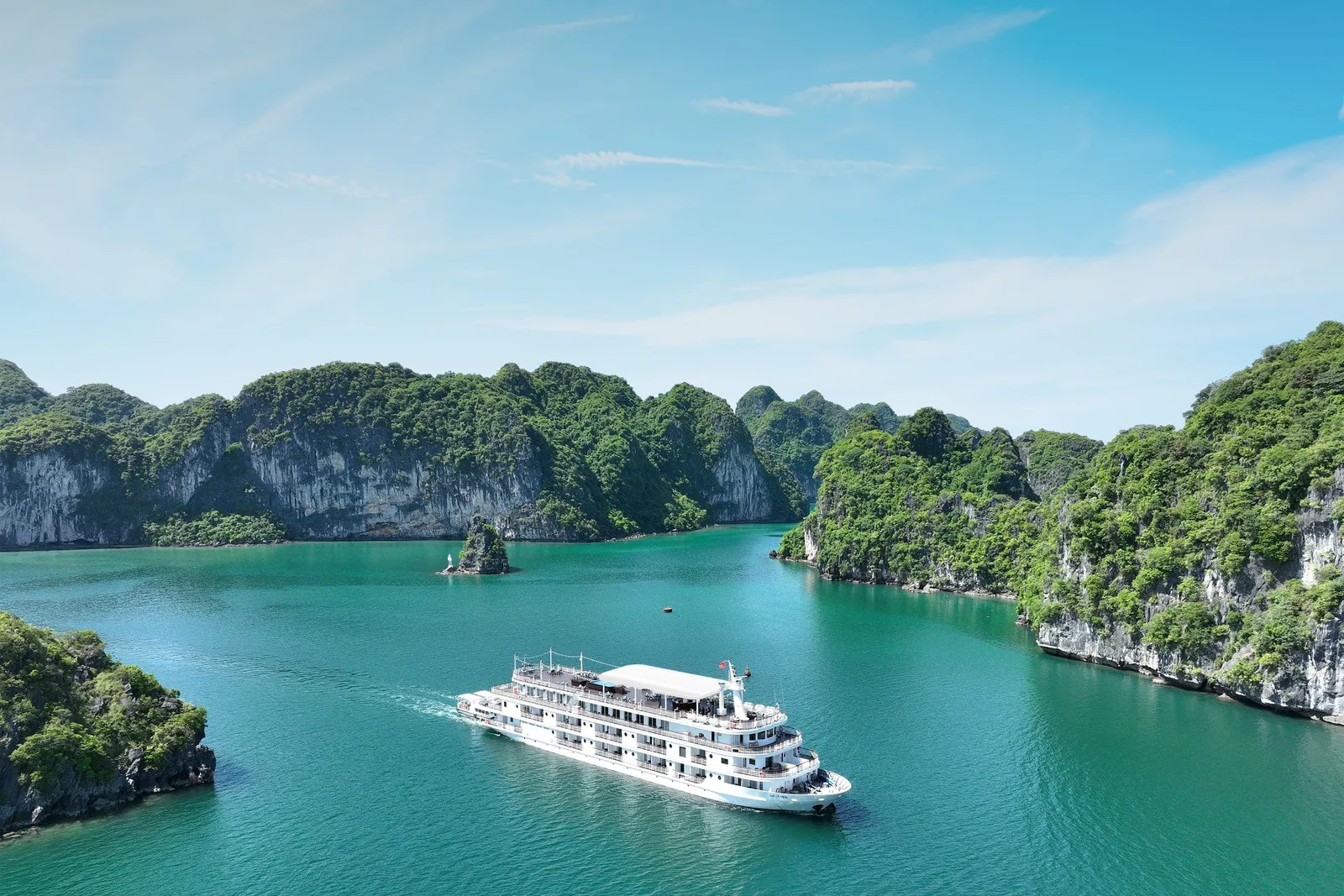 A white multi-deck cruise ship sailing through emerald green water surrounded by limestone karst islands covered in vegetation.