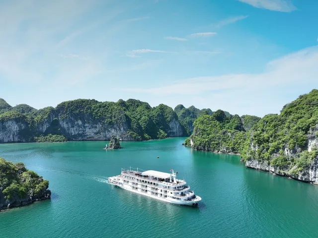 A white multi-deck cruise ship sailing through emerald green water surrounded by limestone karst islands covered in vegetation.