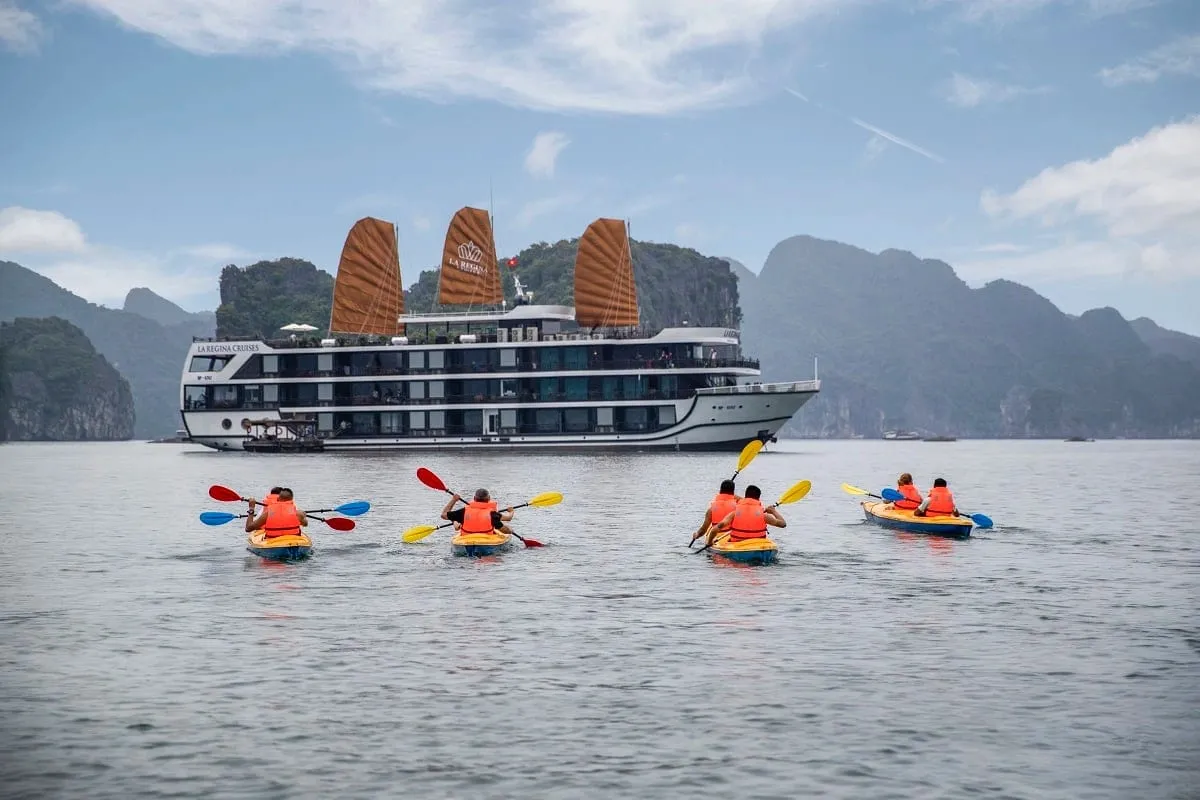 A white multi-deck steel cruise ship with three orange sails sits in a karst bay while several kayakers in orange life vests paddle toward it.