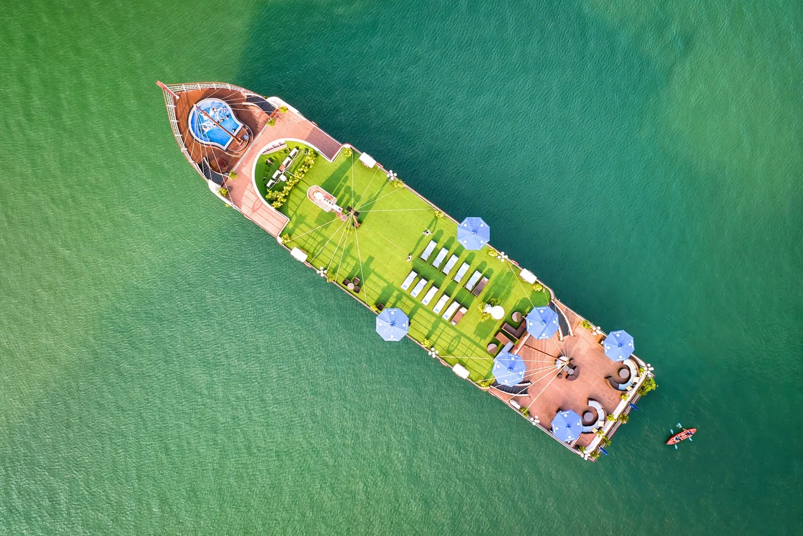 Aerial view of a cruise boat with green artificial turf deck, blue umbrellas, and white lounge chairs floating in turquoise water with a kayaker nearby.