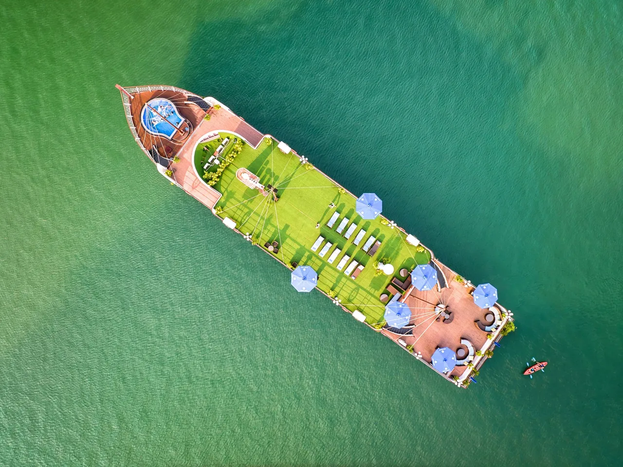 Aerial view of a cruise boat with green artificial turf deck, blue umbrellas, and white lounge chairs floating in turquoise water with a kayaker nearby.